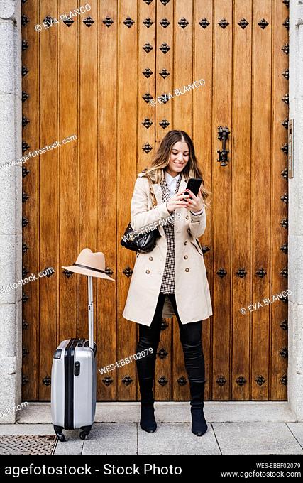 Smiling mid adult woman with wheeled luggage using mobile phone against wood door