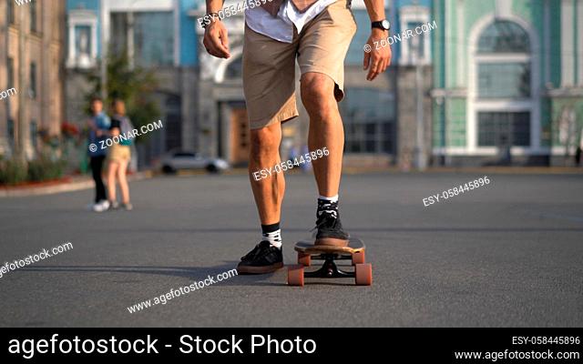 The skater is moving quickly straight ahead on a sunny afternoon. The legs of a light-skinned man are gaining speed on a longboard against the backdrop of city...