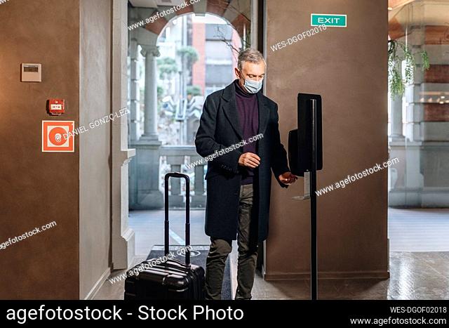 Man with protective face mask applying hand sanitizer while standing by luggage at hotel