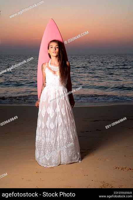 Romantic portrait of beautiful brunette woman in white lace dress standing with pink surfboard at the beach over sea and sunset sky background