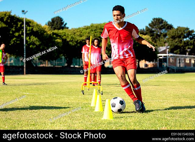 Multiracial male players in red jersey running with soccer ball between cones at playground