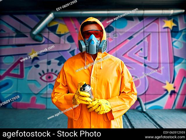 Graffiti painter in respirator mask standing near the wall with his paintings looking to camera. Street art concept