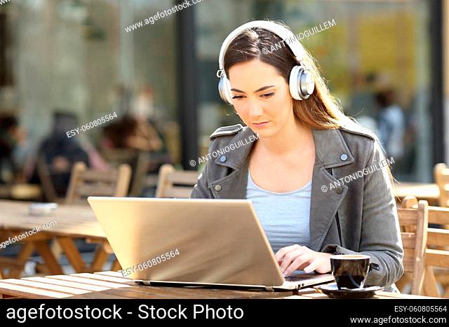 Serious girl e-learning on her laptop with headphones on a coffee shop terrace