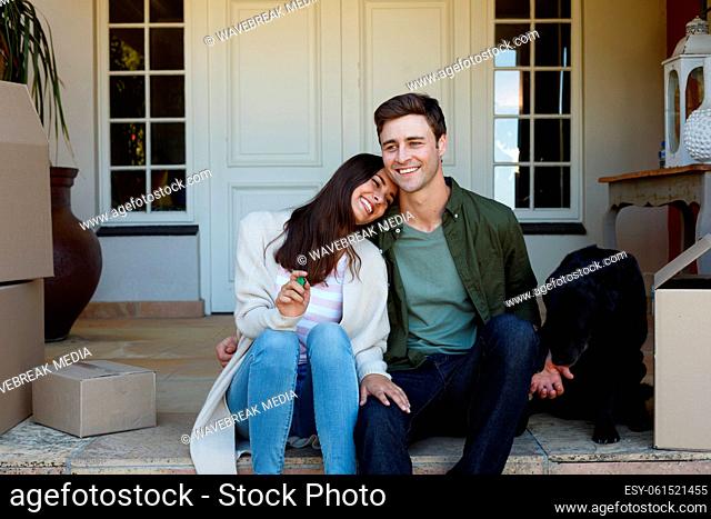 Smiling caucasian couple holding keys sitting outside house with dog