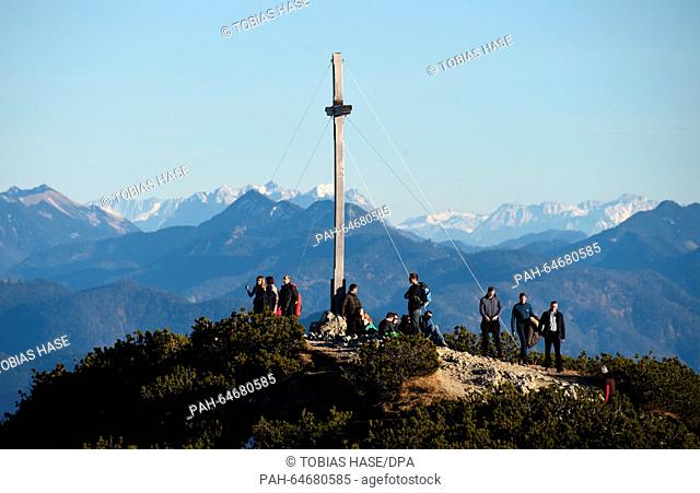 Mountain hikers stand on the viewing plattform next to the summit cross on the mountain peak of the Herzogstand enjoying the warm sun near lake Walchensee