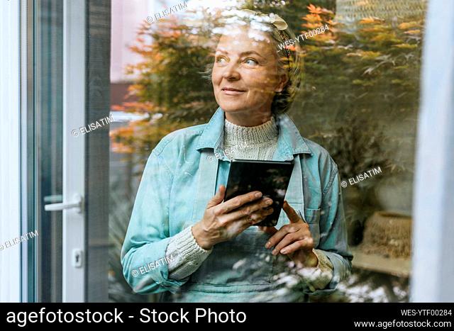 Thoughtful craftswoman holding tablet PC in workshop seen through glass window