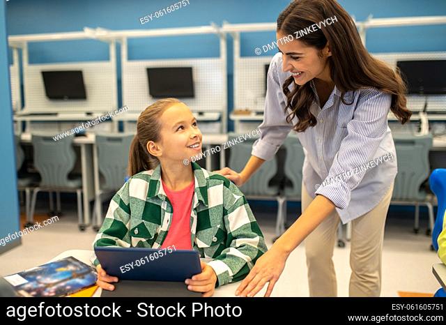 Communication. Girl sitting at desk with tablet and teacher standing leaning smiling looking at each other in class