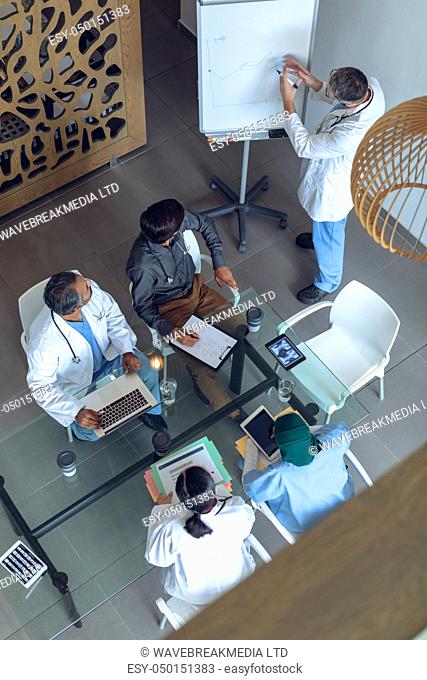 High angle view of Caucasian male doctor explaining graph flip chart in meeting with diverse medical team at hospital . Coffee cup, medical folders, clipboard