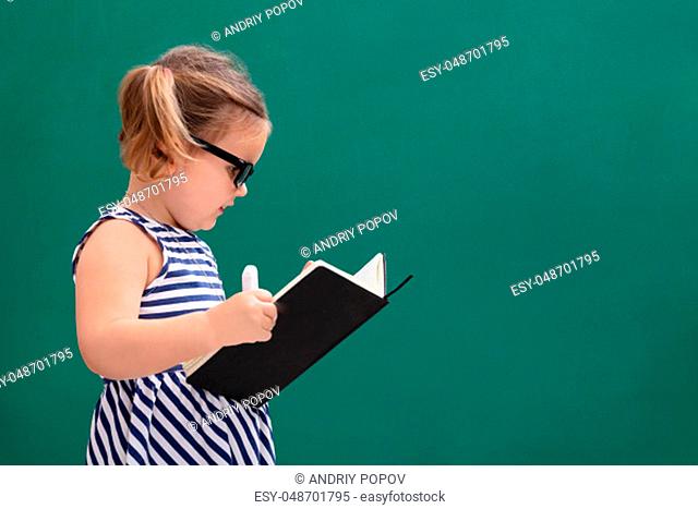 Cute Little Girl Standing In Front Of Chalkboard Reading Book