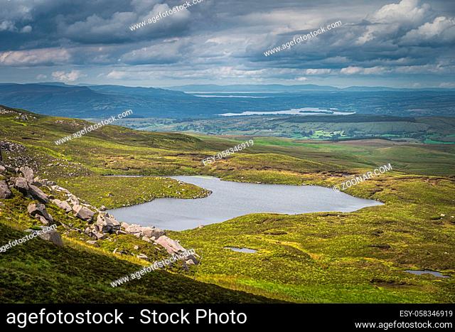 Rolling green hills and fields illuminated by sunlight in Cuilcagh Mountain Park with small lakes and dramatic stormy sky, Northern Ireland