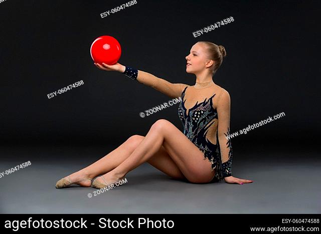 Beautiful blond gymnastist girl in black sparkling costume holding red ball over black background