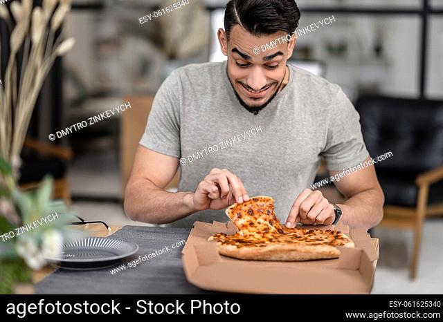 Favorite food. Joyful young bearded man with looking at fresh pizza taking slice sitting at table in apartment