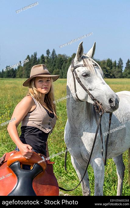 Junge Frau mit Cowboyhut steht mit ihrem Reitsattel vor ihrem Pferd