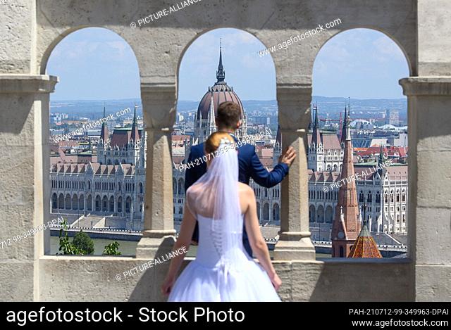 26 June 2021, Hungary, Budapest: A wedding couple stands by the arches of the Fishermen's Bastion with the Parliament building on the Danube in the background
