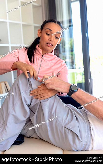 Biracial female physiotherapist giving leg massage therapy to caucasian senior man at home