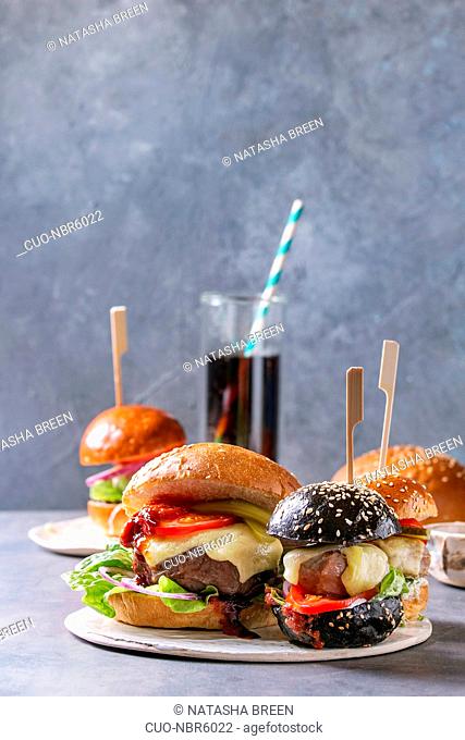 Variety of homemade classic and mini burgers in wheat and black buns with beef and veal cutlets, melted cheese and vegetables on white ceramic board over grey...