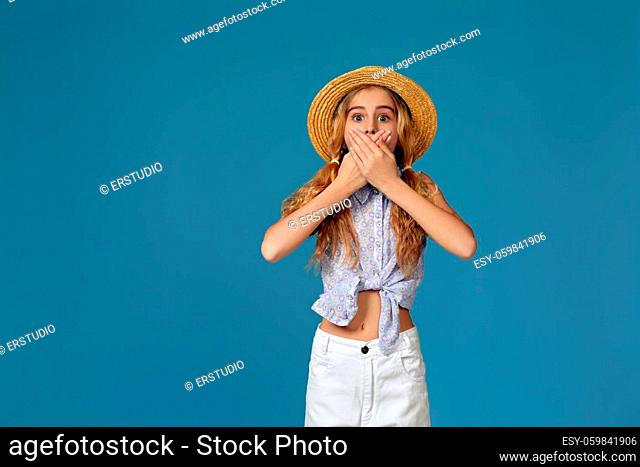 surprised little girl in summer hat covers her mouth with her hands on blue background