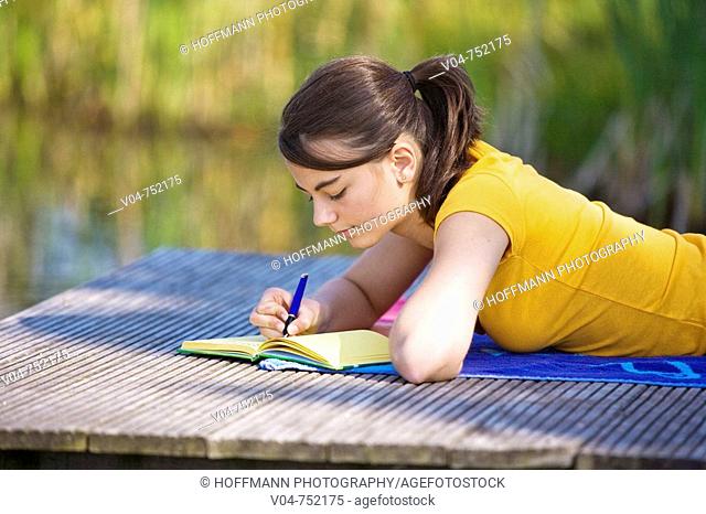 Teenaged girl lying on a footbridge and writing in her diary looking thoughtful
