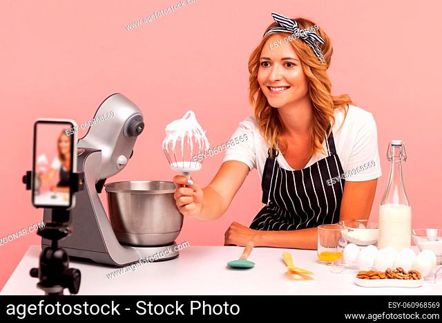 Young adult female food blogger broadcasting process of making pastry, expressing positive emotions, showing whisk with cream to phone on tripod, vlog