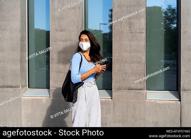 Young woman in protective face mask walking by building during COVID-19