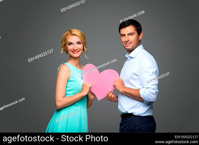 Beautiful young couple holding big pink hearts. Blond woman in mint dress and handsome man in shirt and trousers standing on grey background
