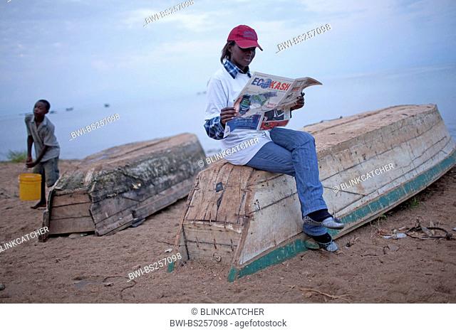 young woman sitting on an upside-down boat on the sand beach reading a paper while a boy is fetching water in a bucket from Lake Tanganijka, Burundi, Nyanza Lac