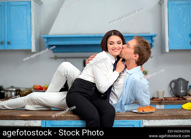 Happy couple higging in kitchen at home. Romantic couple in kitchen sitting on counter. Happy man kissing his gorgeous wife while laying on modern kitchen table