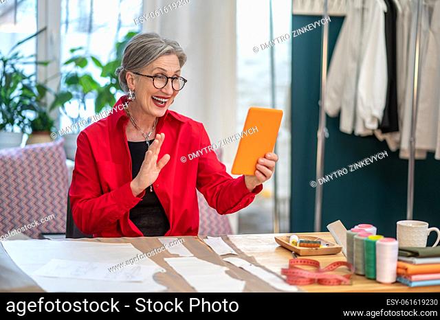 Video call. Joyful gray-haired woman in glasses gesturing greeting looking into smartphone sitting at table in her atelier