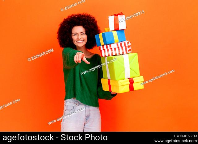 Beautiful woman with curly hair wearing green casual style sweater pointing finger to camera, holding stack of presents, choosing you as winner