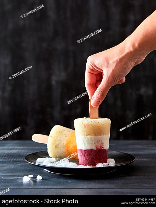 A woman's hand takes a fruity coffee homemade ice cream lolly in a black plate with ice cubes on a dark wooden background with copy space