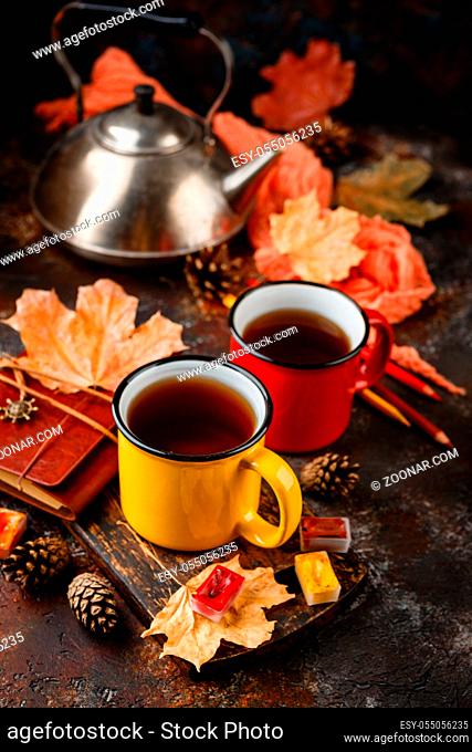Two colored red and yellow enamel cups of tea, watercolors in cuvettes, colored pencils, autumn maple leaves and bumps on a rusty brown background