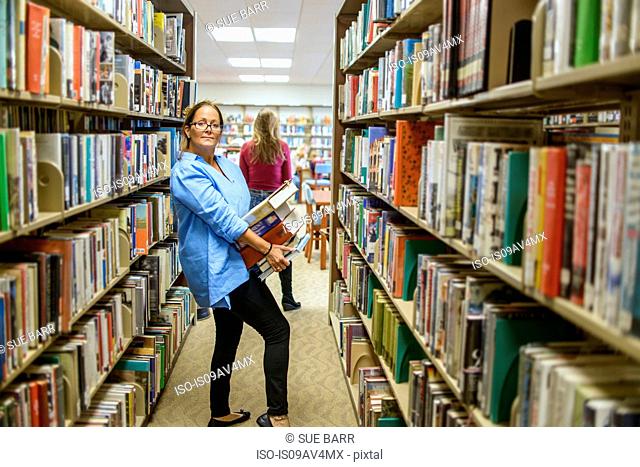 Portrait of mature woman carrying stack of books from library shelves