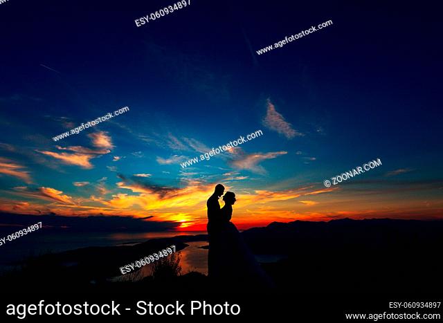 The bride and groom hugging on the top of Mount Lovcen overlooking the Bay of Kotor by the sunset, silhouettes . High quality photo