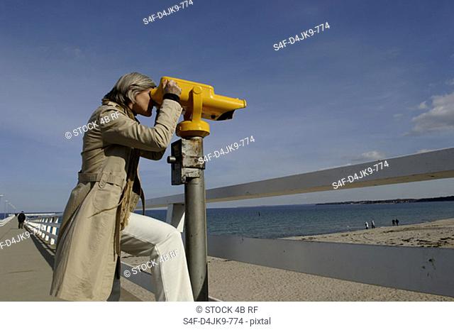 Mature woman looking through a coin-operated binoculars over the Baltic Sea beach