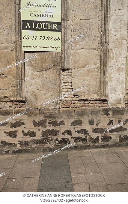 Deteriorating building with crumbling plasterwork revealing brick interior advertised for rent by elegant copperplate sign in the historic centre of Cambrai