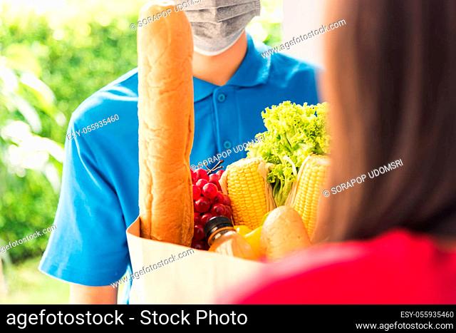 Asian young delivery man in uniform wear protective face mask he making grocery service giving fresh food to woman customer receiving front house under pandemic...