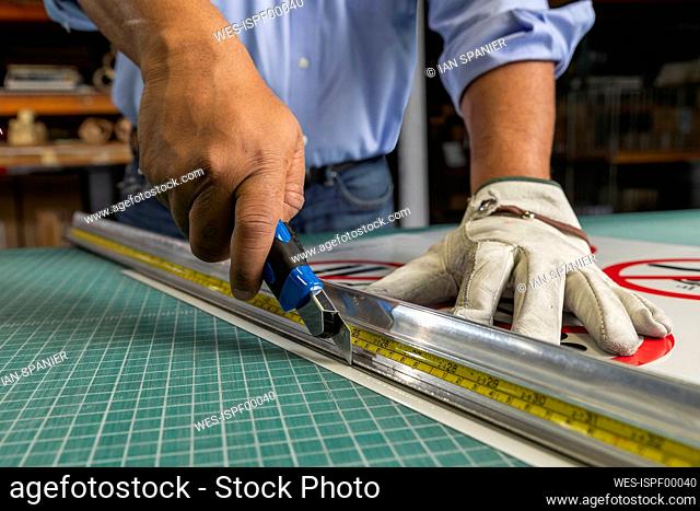 Male employee cutting paper with equipment and ruler on workbench in shop
