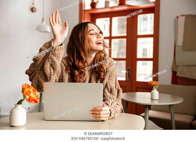 Portrait of a smiling young woman waving her hand while sitting at the table with laptop computer indoors