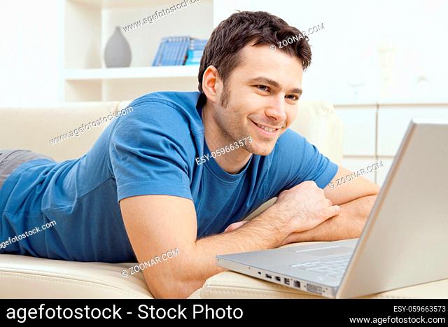 Happy young man laying on sofa and using laptop computer at home, smiling