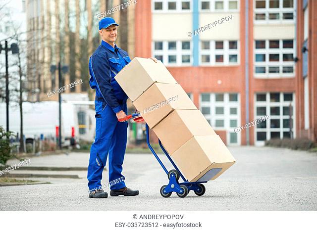 Mature Happy Deliveryman Holding Trolley Loaded With Cardboard Boxes On Street