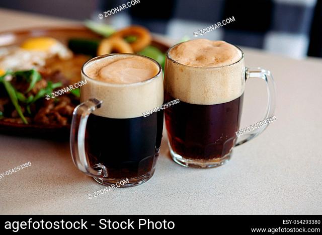 Two big glass cups full of foamy cold beer standing on light smooth table surface in restaurant or pub. Big plate with delicious appetizers like potato puncakes...
