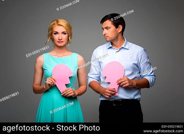Beautiful young couple holding big pink broken heart. Blond woman in mint dress and handsome man in shirt and trousers standing on grey background