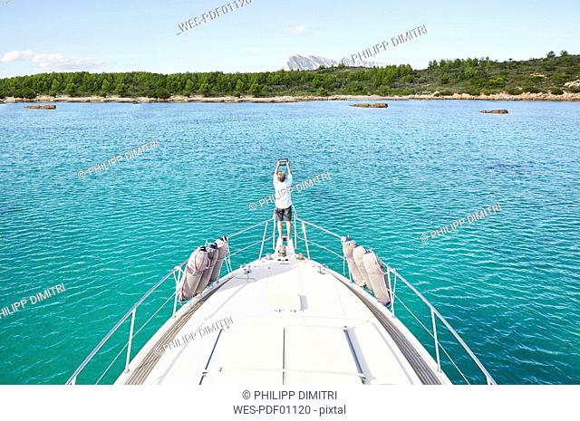 Back view of man standing at bow of his motor yacht taking picture with tablet