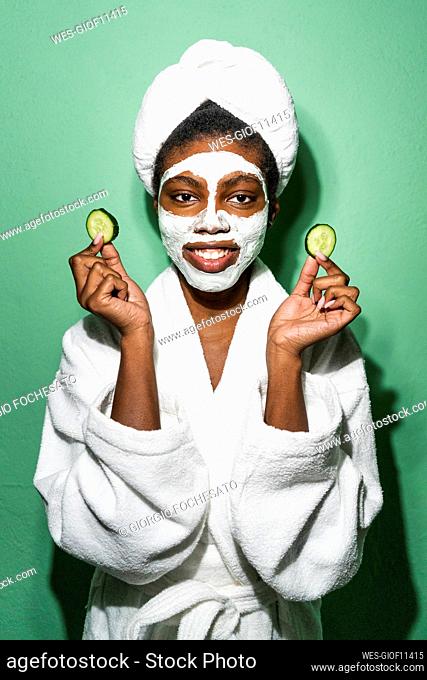 Young woman with facial mask holding cucumber while standing against green background