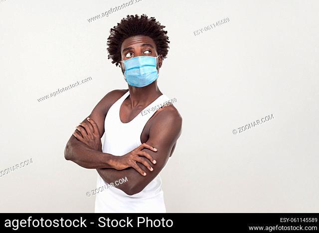 Portrait of thoughtful young man wearing white shirt with surgical medical mask standing, folding arms and looking away and thinking what to do, boring