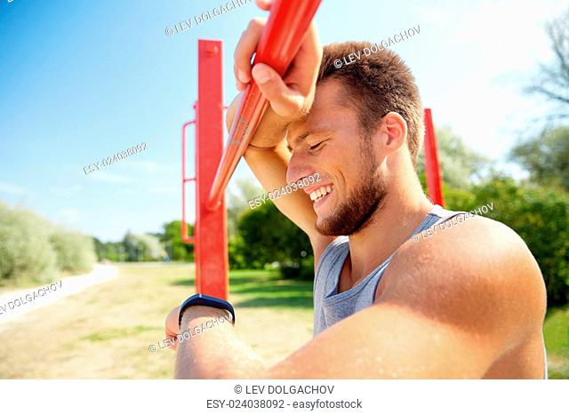 fitness, sport, training and lifestyle concept - happy young man looking at heart-rate watch bracelet and exercising on horizontal bar outdoors