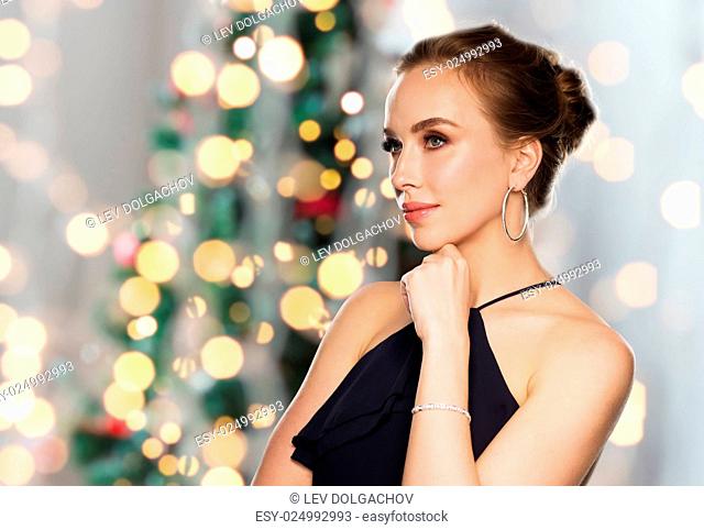 people, luxury, jewelry and holidays concept - beautiful woman in black wearing diamond earrings and bracelet over christmas tree lights background