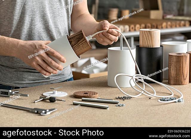 Assembling in the workshop. Man holds a white lamp with a wooden part per the cable. On the table there are other lamp billets, different wrenches, caliber