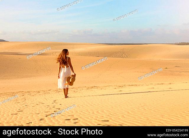 Young beautiful woman with white dress walking in the desert dunes during sunset. Girl walking on golden sand on Corralejo Dunas, Fuerteventura