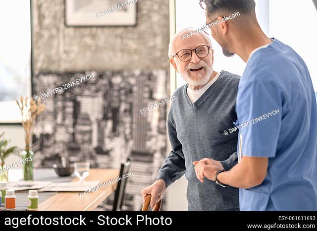 Joyous elderly person in eyeglasses leaning on the walking stick assisted by his in-home caregiver
